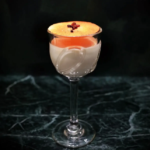 Layered cocktail in a clear stemmed glass with orange foam on top and a small red garnish on a dark marble surface