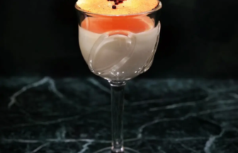 Layered cocktail in a clear stemmed glass with orange foam on top and a small red garnish on a dark marble surface