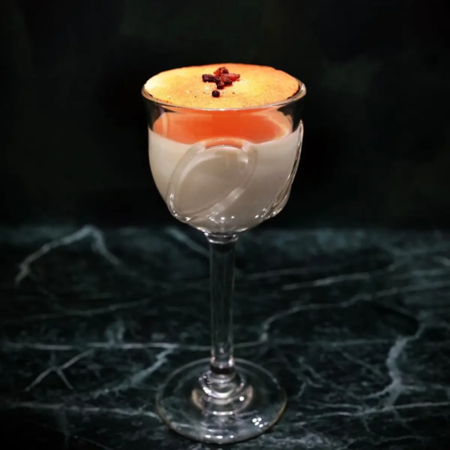 Layered cocktail in a clear stemmed glass with orange foam on top and a small red garnish on a dark marble surface