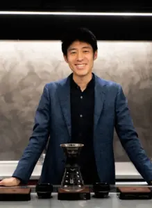 Smiling man in a blue blazer stands behind a pour over coffee setup on a counter with a glass carafe and dripper nearby