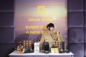 Woman in a beige coat holding a cup behind a coffee product display with capsules and machines on a white table and a city backdrop behind her