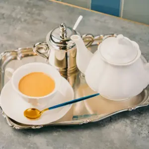 White porcelain teapot teacup with amber tea and a spoon on a polished silver trayclassic tea service setup