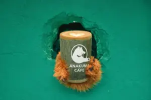 b9dd9565aa480b0e8a8aa5ee3dd91fc2 | コーヒーステーション Latte with a heart latte art in a wooden cup featuring the Anakuma Café logo held by orange furry paws emerging from a green wall crack
