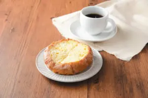 bf64d89011b69b044bb0463fcd22b0a5 | コーヒーステーション Crusty circular loaf on a small plate beside a cup of black coffee on a wooden table unfolded with a beige cloth nearby