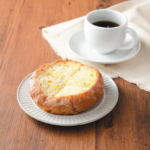 Crusty bread on a white plate beside a cup of black coffee on a wooden table with a light cloth nearby