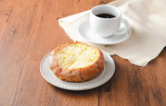 Crusty bread on a white plate beside a cup of black coffee on a wooden table with a light cloth nearby