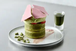 Stack of green matcha pancakes topped with pink brittle shards red filling and green sauce drizzle on a white plate with small crumble bits nearby