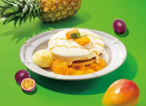 Dessert on a white plate with a glossy white glaze and mango chunks topped with mint on a bright green surface with pineapple and passion fruit nearby