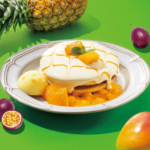 Dessert plate featuring a frosted cake with mango pieces mint and caramel drizzle on a white plate against a bright green background with tropical fruits nearby