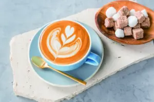 Blue cup of latte with leaf latte art on a light blue saucer with a gold spoon beside a wooden bowl of sugar cubes and marshmallows on a white stone slab