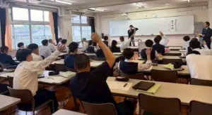 Classroom scene with students in uniforms raising hands as a teacher speaks at the front by a whiteboard with windows behind