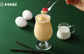 Creamy dessert drink in a tall glass topped with vanilla ice cream and a cherry on a spoon, with a milk jug, eggs, and vanilla sticks on a green background.