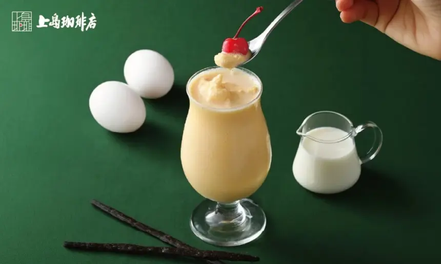 Creamy dessert drink in a tall glass topped with vanilla ice cream and a cherry on a spoon with a milk jug eggs and vanilla sticks on a green background