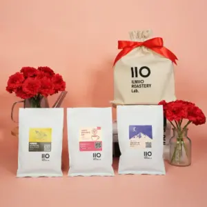 cf3b2b1bec95cc50b0d77b1d8e76dd87 | コーヒーステーション Display of three white coffee bags with colorful labels in front of a large cream gift bag tied with a red ribbon pink background and red carnations behind