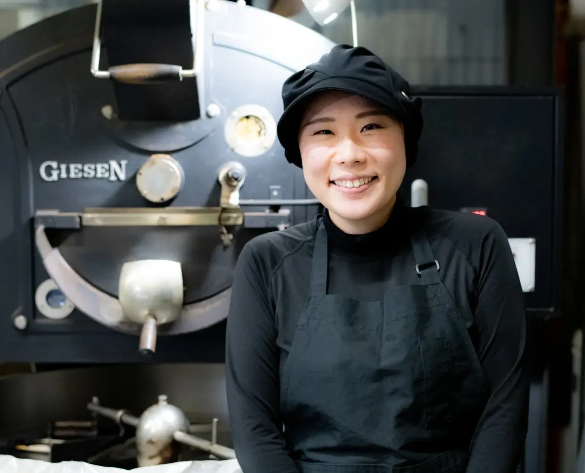 コーヒー焙煎機の前で笑顔を見せる女性のポートレートです。黒いエプロンと帽子を着用しており、コーヒー店員または焙煎士のようです。背景にはGIESEN製の焙煎機が見えます。