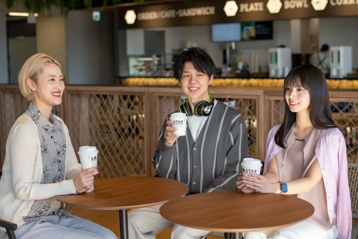 明るいカフェまたはフードコートで、3人の若者（女性2人、男性1人）がコーヒーカップを手に笑顔で会話を楽しんでいる様子。休憩時間やランチタイムを一緒に過ごしているようです。