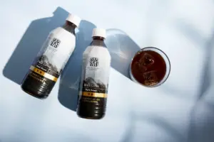 Two bottles of black coffee labeled Dark Roast beside a glass of iced coffee on a pale blue background