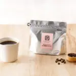 Cup of black coffee beside a silver resealable coffee bag with pink label and a wooden scoop of beans on a light wood table