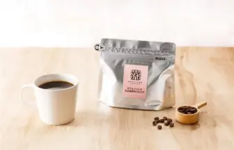 Cup of black coffee beside a silver resealable coffee bag with pink label and a wooden scoop of beans on a light wood table