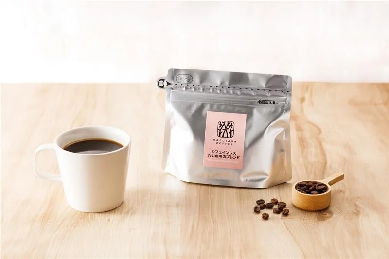 Cup of black coffee beside a silver resealable coffee bag with pink label and a wooden scoop of beans on a light wood table