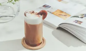 Tall glass of iced latte with foamy top and a cinnamon garnish on a wooden coaster beside an open magazine on a bright table