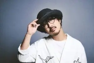Young man wearing a black fedora tilting his hat with one hand in a white shirt against a gray studio background