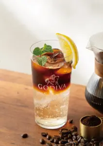 Tall glass of iced beverage branded Godiva Café with lemon wedge mint and ice on a wooden table