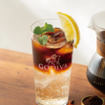Tall glass of iced Godiva chocolate coffee cocktail with a lemon wedge and mint garnish on a wooden table with coffee beans and a carafe nearby