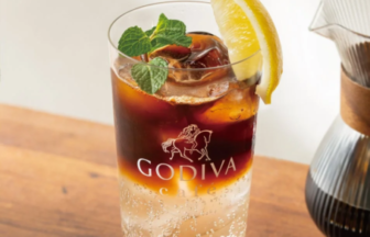 Tall glass of iced Godiva chocolate coffee cocktail with a lemon wedge and mint garnish on a wooden table, with coffee beans and a carafe nearby.