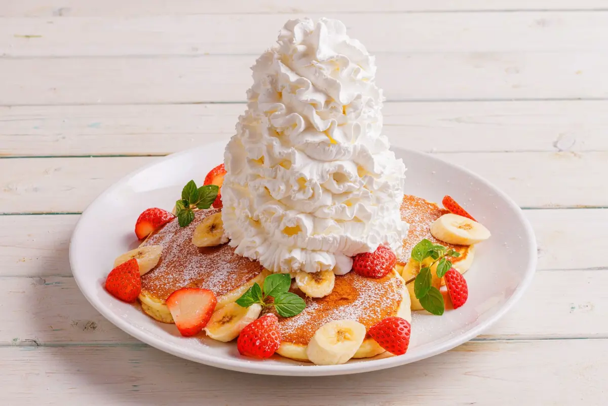 白いお皿に盛られた、たっぷりのホイップクリームとイチゴ、バナナ、ミントが飾られたパンケーキの画像です。粉砂糖が振りかけられ、木製のテーブルに置かれています。