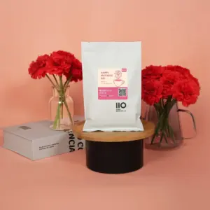 e910f8897902d3c4ae74e520508fe21a | コーヒーステーション White coffee bag with a pink label reading Happy Mothers Day on a round wooden stand framed by red carnations in glass vases against a pink background