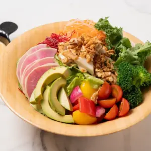 Colorful veggie salad in a wooden bowl with avocado slices radishes cherry tomatoes broccoli kale shredded carrots beets and walnuts topped with crispy onions and greens great for a healthy meal