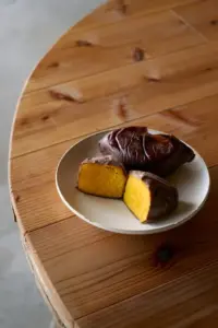 Two roasted purple skin sweet potatoes on a white plate one cut open to reveal bright orange flesh set on a wooden table