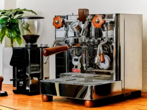 Shiny espresso machine with wooden handles and gauges on a wooden counter next to a coffee grinder and plant in the background