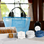 Blue gift bag with assorted coffee and chocolate gift set displayed on a white table in a sunlit room includes cookies tins and a liquid coffee bottle