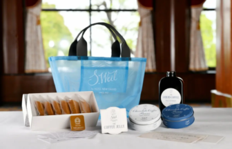 Blue gift bag with assorted coffee and chocolate gift set displayed on a white table in a sunlit room, includes cookies, tins, and a liquid coffee bottle.