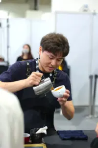 Barista pours espresso from a metal jug into a white cup at a cafe setup focused on the task