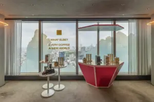 Promotional showroom with gold coffee product displays on white tiered stands and a pink angular counter overlooking a city skyline text reads WHAT ELSE EVERY COFFEE A NEW WORLD