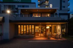 Nighttime exterior of a modern restaurant with warm interior lighting floor to ceiling windows and outdoor seating on the right a glowing S sign is visible