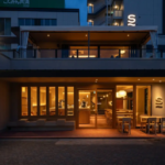 Exterior of a warmly lit cafe at dusk with large glass windows and a second floor terrace Outdoor seating is on the right side of the entrance