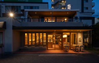 Exterior of a warmly lit cafe at dusk with large glass windows and a second-floor terrace. Outdoor seating is on the right side of the entrance.