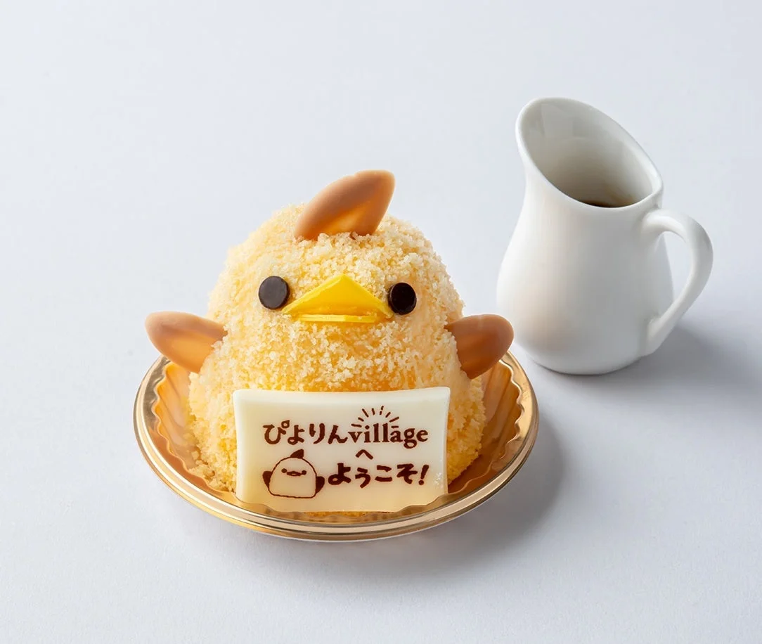 白い背景に、ひよこの形をした可愛らしいケーキが置かれています。ケーキの手前には「ぴよりんvillage へ ようこそ！」と書かれた白いチョコレートプレートがあり、隣には白い小さなピッチャーが添えられています。