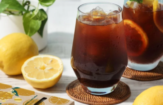 Two glasses of iced tea with ice and lemon wedges sit on a white table, surrounded by lemons, a spoonful of brown sugar, and a potted plant.
