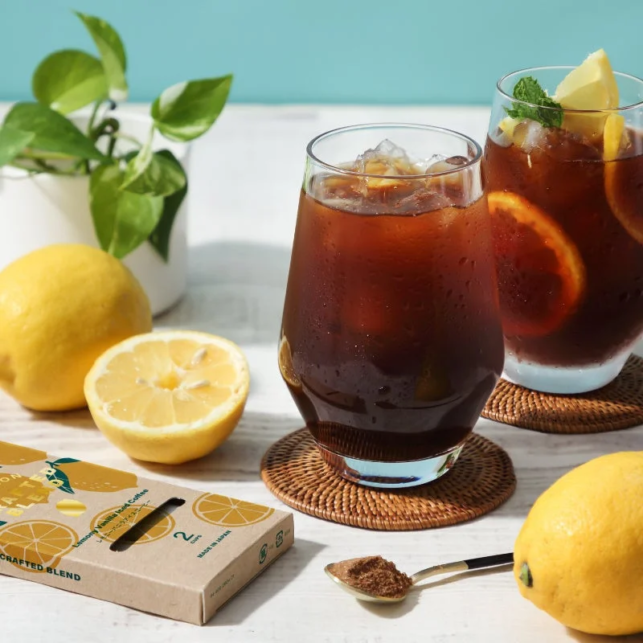 Two glasses of iced tea with ice and lemon wedges sit on a white table surrounded by lemons a spoonful of brown sugar and a potted plant
