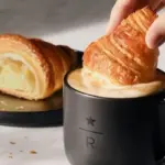 Croissant being dipped into a black mug of coffee while another croissant rests on a plate beside it
