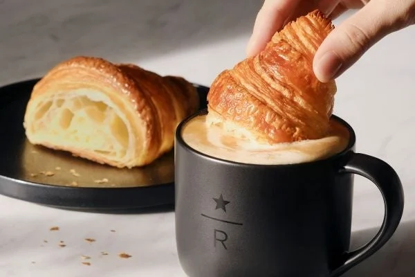 Croissant being dipped into a black mug of coffee while another croissant rests on a plate beside it