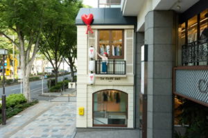 Two story storefront on a tree lined street with a mannequin on the balcony holding a red heart balloon above the sign Le Café Ami