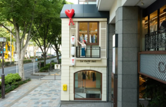 Two story storefront on a tree lined street with a mannequin on the balcony holding a red heart balloon above the sign Le Café Ami