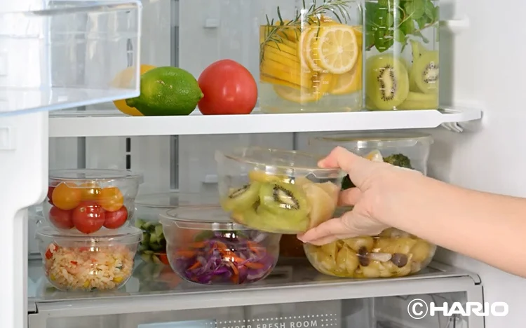 Person places a clear plastic container of sliced kiwi into a neatly organized fridge with prepped fruit and vegetables in containers on shelves