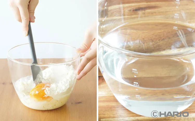 Person mixing egg and flour in a glass bowl with a spatula on a wooden table next to it a clear measuring beaker with water on wood